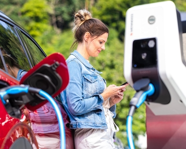 Eine Frau lehnt an ihr Elektroauto und drückt auf ihrem Smartphone herum, während das Auto mit einer Ladesäule geladen wird.